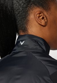 Černá bunda s vysokým límcem. Límec má reflexní stříbrné logo. Hladká textura s kontrastním matným a lesklým povrchem.