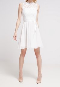 Vit ärmlös klänning med en spetsöverdel och en utsvängd chiffongkjol, med ett bälte av tyg i midjan och en slät textur.