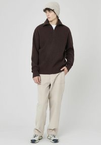 Pull en maille marron avec un col zippé, assorti à un pantalon texturé beige clair et un bonnet gris clair. Des baskets complètent le look.