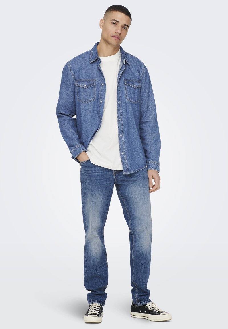 Denim košulja dugih rukava u srednje plavoj boji, s dva džepa na prsima i klasičnom kragnom. Uparena s laganim farmerkama i crnim tenisicama.