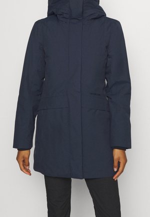 Personne portant un long manteau d'hiver bleu marine avec un col montant et une capuche, debout devant un fond clair uni.
