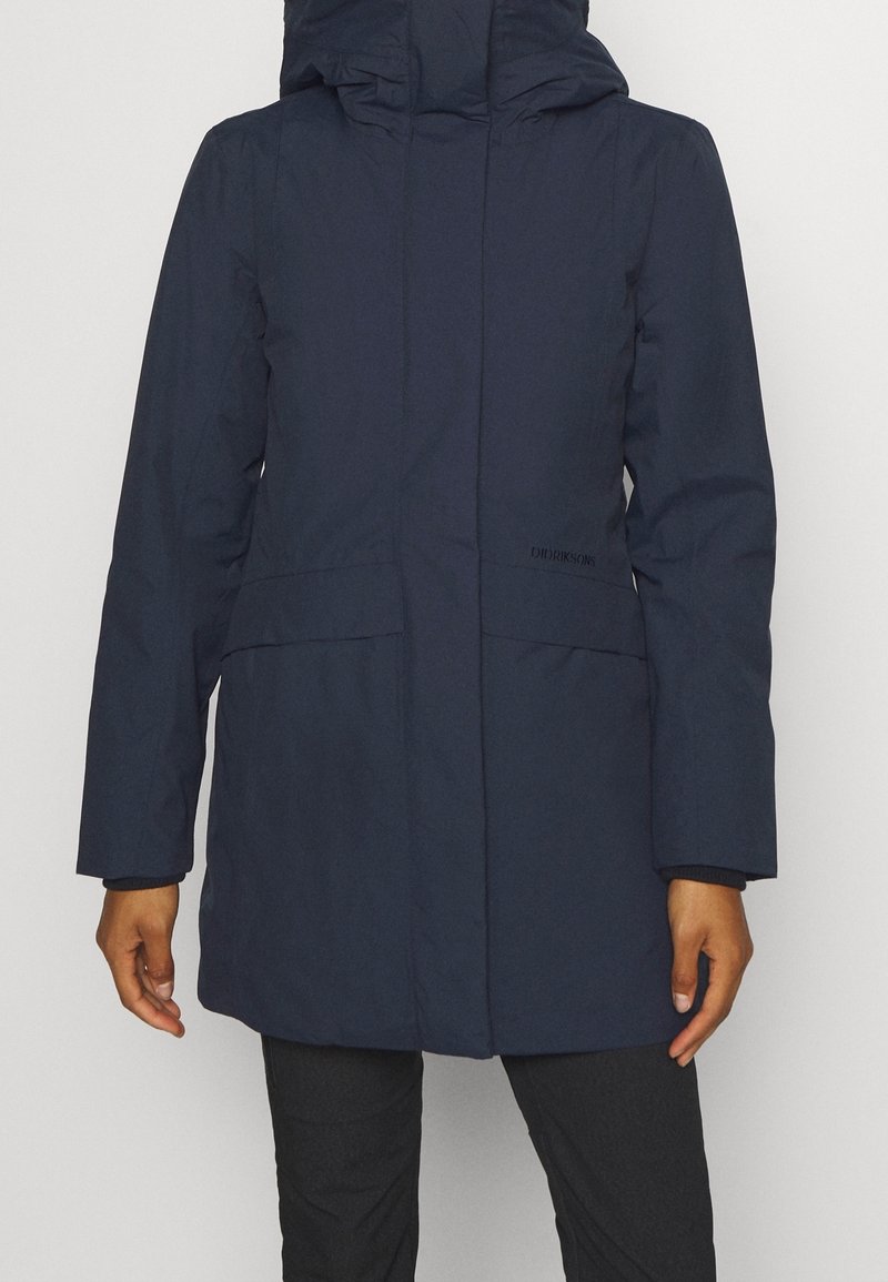 Personne portant un long manteau d'hiver bleu marine avec un col montant et une capuche, debout devant un fond clair uni.