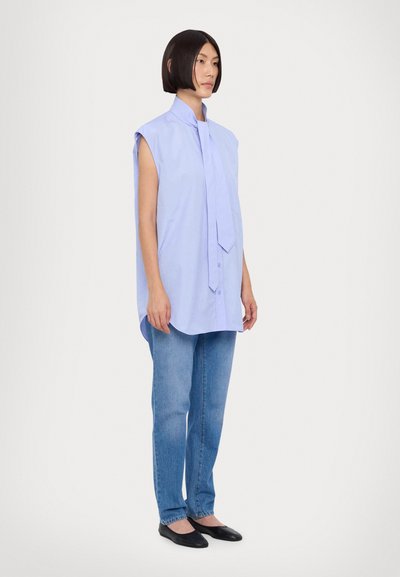 Maglietta senza maniche di un delicato colore viola chiaro con colletto alto e cravatta, abbinata a jeans azzurri e ballerine nere. Tessuto liscio, vestibilità comoda.