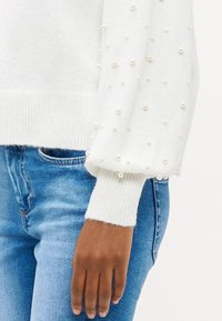 Biały sweter z fakturowanym splotem, z bufkami ozdobionymi rozsypanymi sztucznymi perłami. W połączeniu z dopasowanymi niebieskimi dżinsami.