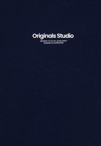 Tamno plava pamučna majica s bijelim natpisom "Originals Studio" i "Brought to you by JACK&JONES, Designed in Copenhagen" iznad.