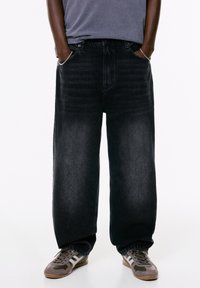 Person iført løse sorte jeans med hænderne i lommerne, grå t-shirt og grå-hvide sneakers foran en hvid baggrund.