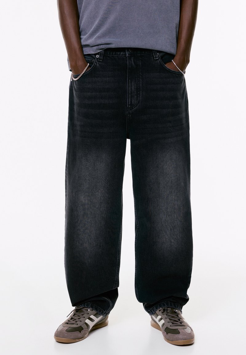 Person, der weite schwarze Jeans trägt, mit Händen in den Taschen, grauem T-Shirt und grauen und weißen Sneakers vor einem weißen Hintergrund.