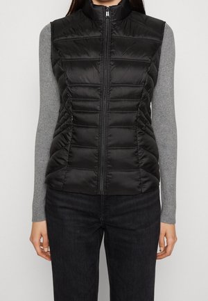 Woman wearing a black quilted sleeveless vest over a grey long-sleeve top and black pants, standing against a plain background.