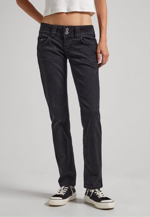 Personne portant un jean noir slim avec fermeture à double bouton et des baskets noires à lacets blancs, debout devant un fond uni.