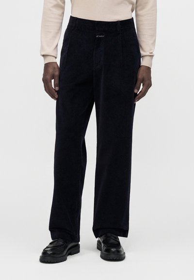 Pantalon large en velours côtelé noir porté avec des mocassins en cuir noir et une chemise beige à manches longues, montré sur une personne debout.