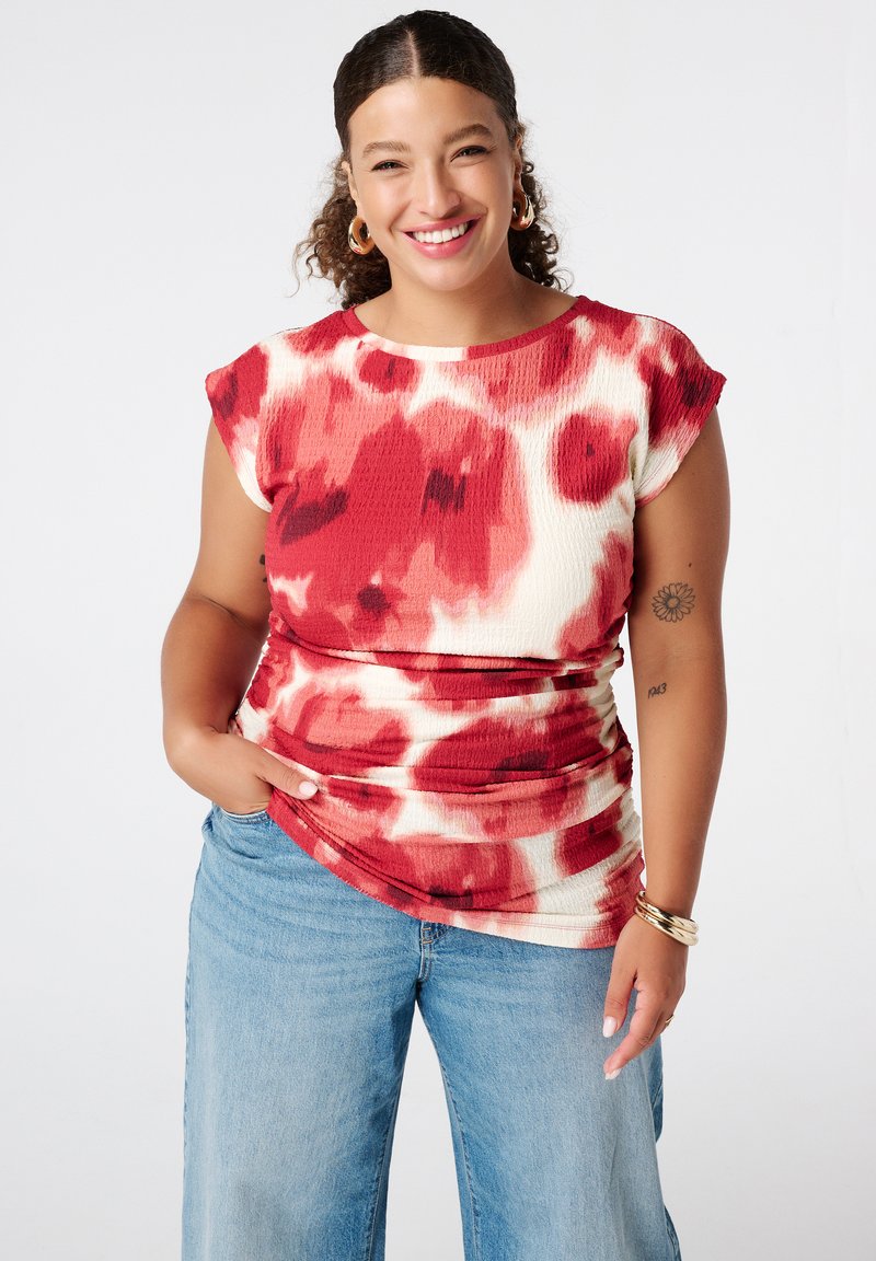 Rood en crème gestippelde top met korte cap sleeves en een textuurafwerking, gecombineerd met lichtblauwe wijde jeans. Kenmerkt ruches bij de taille.