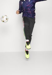 Violetti urheilupaita abstraktilla kuviolla, mustat verkkarit monivärisillä sivuraitoja, ja kirkkaan keltaiset jalkapallokengät.