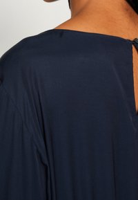 Detailní záběr na horní část zad osoby v tmavě modrém oděvu s kulatým výstřihem a malým zapínáním na knoflík.