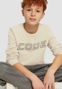 Beige Langarm-T-Shirt mit "CODE" in Dunkelgrau bedruckt, kombiniert mit grauen Hosen. Einfaches Design, weicher Stoff, lässige Passform.