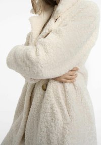 Cappotto color crema, con tessuto strutturato e morbido; presenta un colletto a risvolto e un dettaglio con bottoni sulla parte frontale.