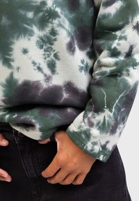 Long-sleeve green and white tie-dye shirt with a fitted cuff and ribbed texture, paired with dark jeans, hands visible at the waist.