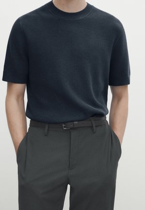 Homme portant un pull en maille bleu marine foncé à manches courtes, rentré dans un pantalon taille haute gris foncé avec une ceinture noire, les mains dans les poches.
