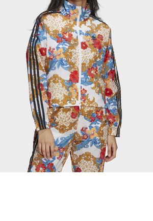 Femme portant un survêtement blanc Adidas avec des motifs floraux et baroques rouges, bleus et dorés et des détails noirs à trois bandes sur les manches et le pantalon.