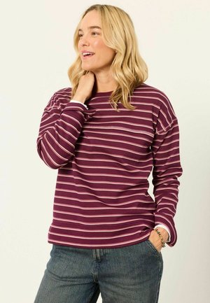 Lyshåret kvinde smiler, iført en langærmet burgundy- og hvidstribet skjorte med blå jeans, den ene hånd rører ved nakken, den anden i lommen.