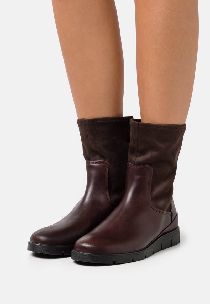 Bottes basses en cuir et suède marron avec une semelle noire en caoutchouc flexible, présentant un design avec revers et une finition lisse et texturée.