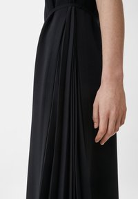 Lähetä lähikuva kädestä, joka levittää mustan laskostetun mekon viereen, jossa on istuva vyötärö ja sileä kangas.