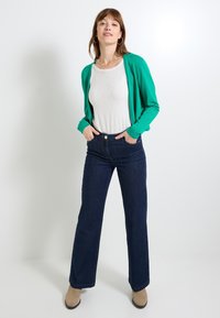 Cárdigan verde sobre una blusa blanca de canalé, combinado con unos vaqueros de campana azul oscuro. La modelo está de pie con las manos en los bolsillos y lleva botas beige.