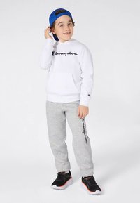 Niño vestido con una sudadera blanca de Champion, pantalones de chándal grises con logo, zapatillas negras con acentos naranjas y una gorra azul puesta al revés, de pie sobre un fondo blanco.