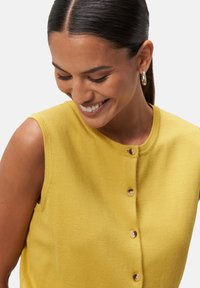 Débardeur en maille jaune sans manches avec un col rond et cinq boutons marron clair. Le tissu a une texture lisse et une silhouette ajustée.