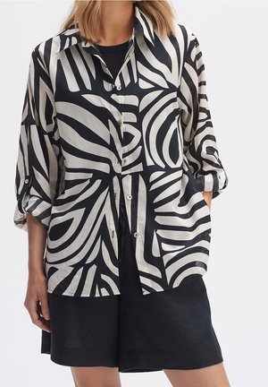 Zwart-wit overhemd met knopen en een patroon van grote, vloeiende abstracte ontwerpen, lange mouwen en een ontspannen pasvorm. Gecombineerd met een donkere korte broek.