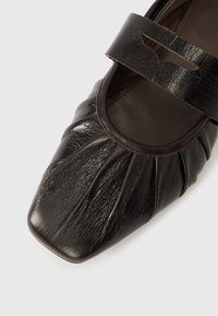 Fekete bőrcipő négyzet alakú orral, redőzött mintával és díszítő pánttal a lábfej fölött. Texturált felület sima részletekkel.