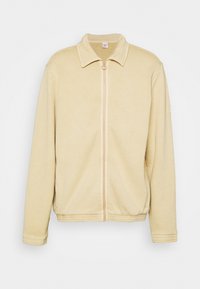 Gul jacka med dragkedja och krage, mjukt, texturerat tyg och resår i midjan. Har ribbstickade muddar på de långa ärmarna.