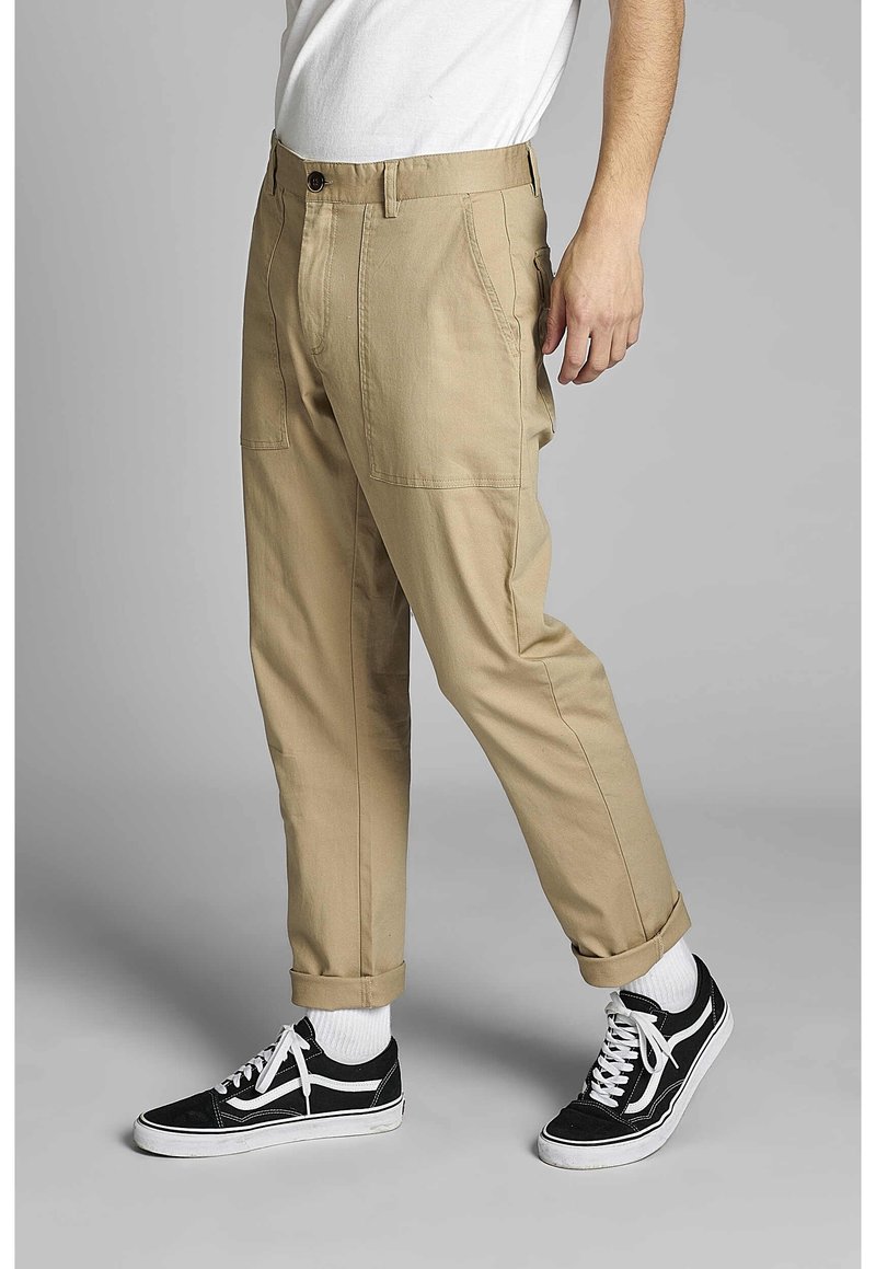 Béžové chino kalhoty s rovný fit, se zadními kapsami a ohrnutými manžetami. Kombinované s černými teniskami a bílými sportovními ponožkami.