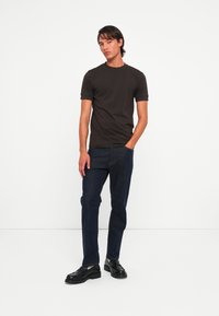 Ung mand står med den ene hånd i lommen, iført en mørkebrun kortærmet skjorte, mørke jeans og sorte lædersko på en hvid baggrund.