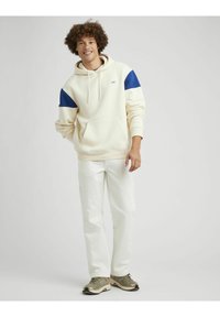 Hoodie crème avec des accents bleus aux épaules, poche kangourou, pantalon blanc et baskets vert olive. Le mannequin se tient devant un fond neutre.
