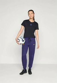 Young woman standing holding a soccer ball in her left hand, wearing a black t-shirt, purple Nike sweatpants, and black sneakers.