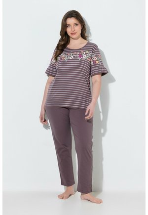 Frau steht barfuß, trägt ein lila gestreiftes Oberteil mit Blumenmuster und passende lila Hose vor weißem Hintergrund.