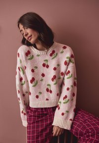Maglione rosa lavorato a maglia con motivi floreali rossi e foglie verdi, caratterizzato da scollo e polsini a coste. Abbinato a una gonna a quadri rossa.