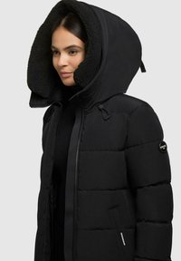 Giubbotto imbottito nero con un grande cappuccio foderato di pelliccia, chiusura con zip, tasche laterali e un patch con logo sulla manica.