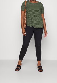 Kvinna iklädd en grönmönstrad kortärmad topp, svarta tajta byxor, svarta klacksandaler och bärande en beige nätad axelväska.