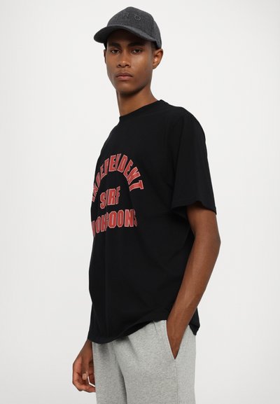 Black cotton t-shirt with red graphic text, rounded neckline, short sleeves, paired with light gray sweatpants and a dark gray cap.