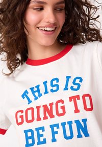 Maglietta bianca a maniche corte con colletto rosso. Testo in grassetto blu e rosso che recita: "QUESTO SARÀ DIVERTENTE." Tessuto di cotone liscio.