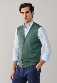 Młody mężczyzna o ciemnych włosach, ubrany w zieloną kamizelkę na guziki, nałożoną na jasnoniebieską koszulę w paski z podwiniętymi rękawami oraz granatowe spodnie, stojący.