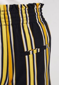 Gros plan sur un pantalon à taille élastique avec des rayures verticales jaunes, noires, blanches et bleues et une petite poche horizontale fendue sur le côté.