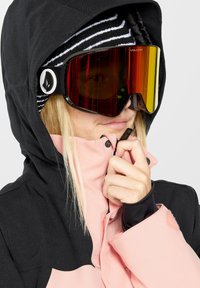 Zwarte en roze sneeuwjas met een hoge kraag en rits, gedragen met een gestreepte beanie en reflecterende skibril met een groot glas.