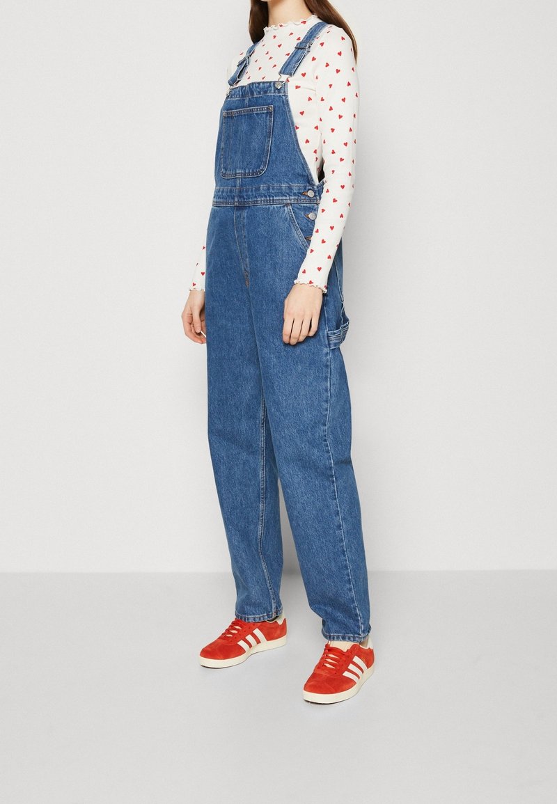 Salopette di denim di colore azzurro chiaro, con tasca frontale, spalline regolabili e gambe larghe. Abbinata a una camicia bianca con cuori rossi e sneaker rosse.