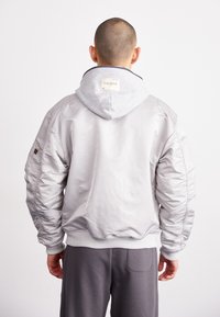 Veste bomber grise avec une finition brillante, dotée d'une capuche grise, de poignets côtelés et d'une étiquette au dos du cou. Texture lisse et coupe décontractée.