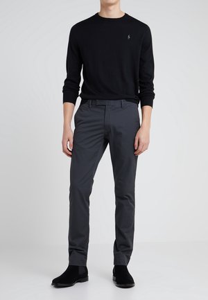 Homme portant une chemise noire à manches longues, un pantalon gris foncé et des bottines en daim noir, debout devant un fond clair uni.