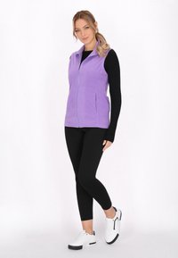 Gilet in pile viola con zip frontale, tasche laterali e colletto. Abbinato a leggings neri e sneakers bianche. Texture liscia.