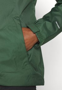 Giacca impermeabile verde con tasca con zip e polsini regolabili. Il materiale ha una texture liscia con una finitura opaca. Accentuata dal logo "DRYVENT".