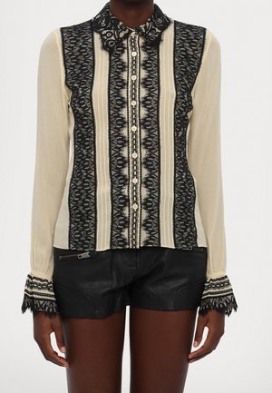Beige Bluse mit langen Ärmeln mit schwarzen vertikalen Spitzenstreifen, spitzenbesetzten Manschetten und Knopfleiste vorne, kombiniert mit schwarzen Leder-Shorts.